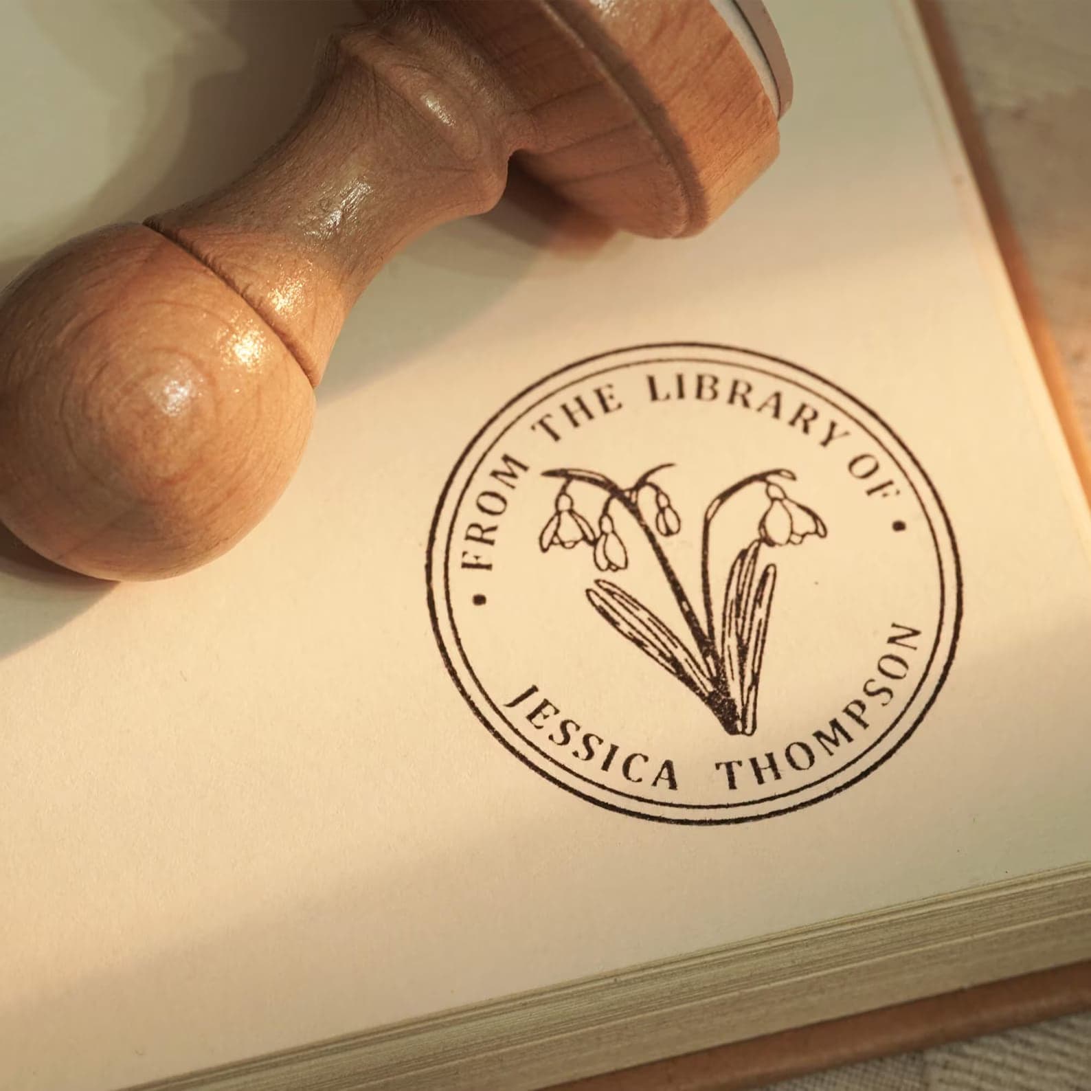 Wooden personalised book ownership stamp beside a printed 'From the library of' ink stamp with a snowdrop design on a book page